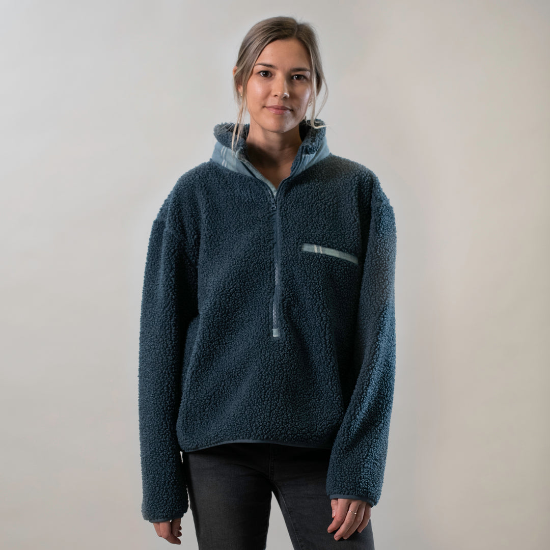 A woman with light brown hair tied back wears a Women's Jenner Sherpa Fleece in Atlantic Blue and dark jeans, standing against a plain light background and looking at the camera with a slight smile.