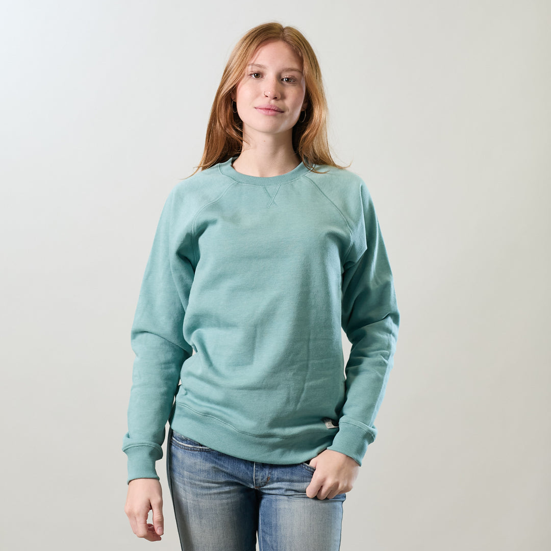 A young woman with long, light brown hair stands against a plain background, wearing the Women's Crew Neck Sweatshirt in Bermuda Green Heather and blue jeans, looking at the camera with a slight smile.