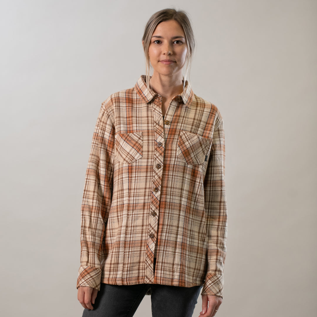 A woman with light brown hair tied back faces the camera, wearing the Women's Fireside Flannel - Cattail Tan with two chest pockets and black pants, standing against a plain light background.