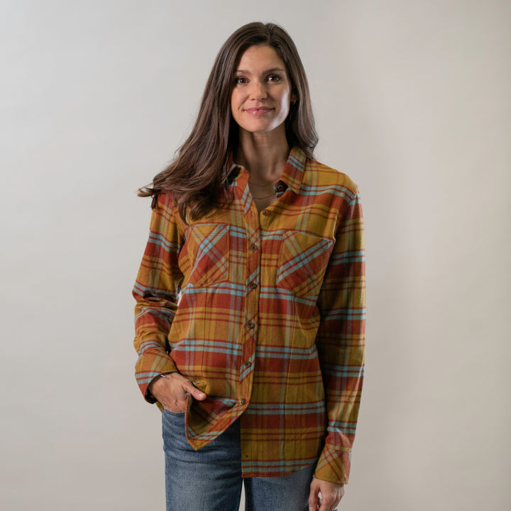 A woman with long brown hair stands against a plain light background, smiling slightly with one hand in her pocket while wearing the Women&