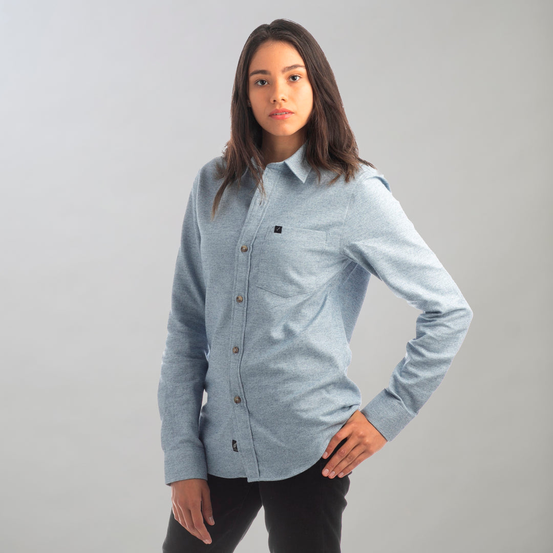 A woman with long dark hair stands against a plain light background, wearing the Women's Cascade Flannel Shirt in Dusty Blue and black pants, one hand on her hip and a neutral expression.