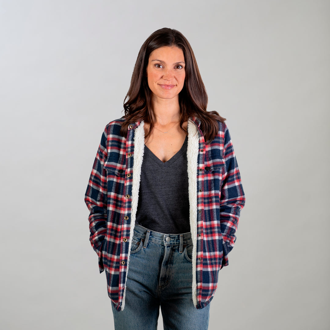 A woman with long brown hair stands against a plain background, wearing the Women's Northwood Sherpa Insulated Flannel Jacket in Eagle Blue over a dark gray V-neck shirt and blue jeans, her hands in her pockets and a slight smile on her face.
