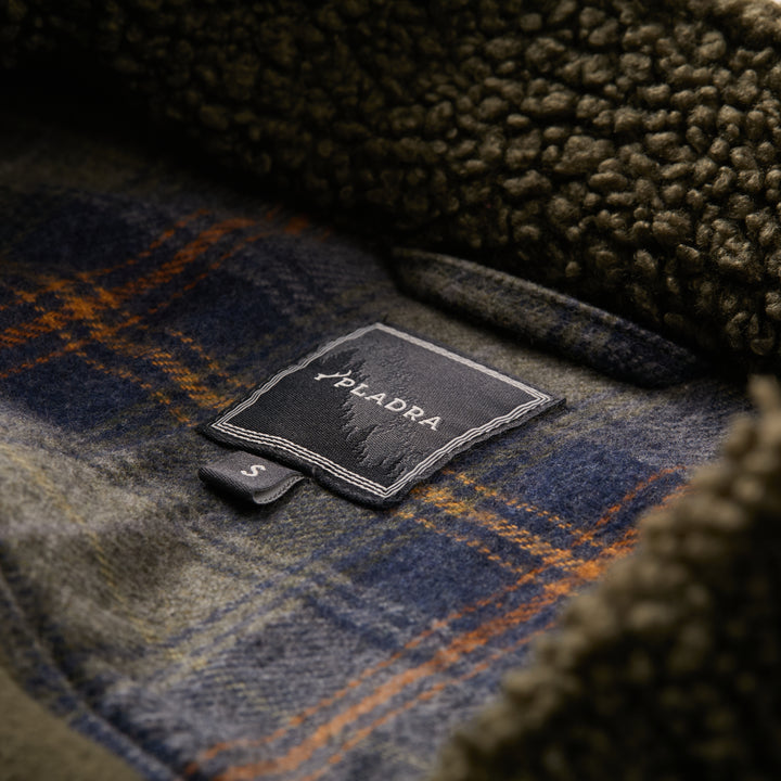 Close-up of the PLADRA S label on blue and green plaid, surrounded by brown recycled polyester fleece—showcasing the cozy details of the Men's Cypress Sherpa Fleece in Forest Green.
