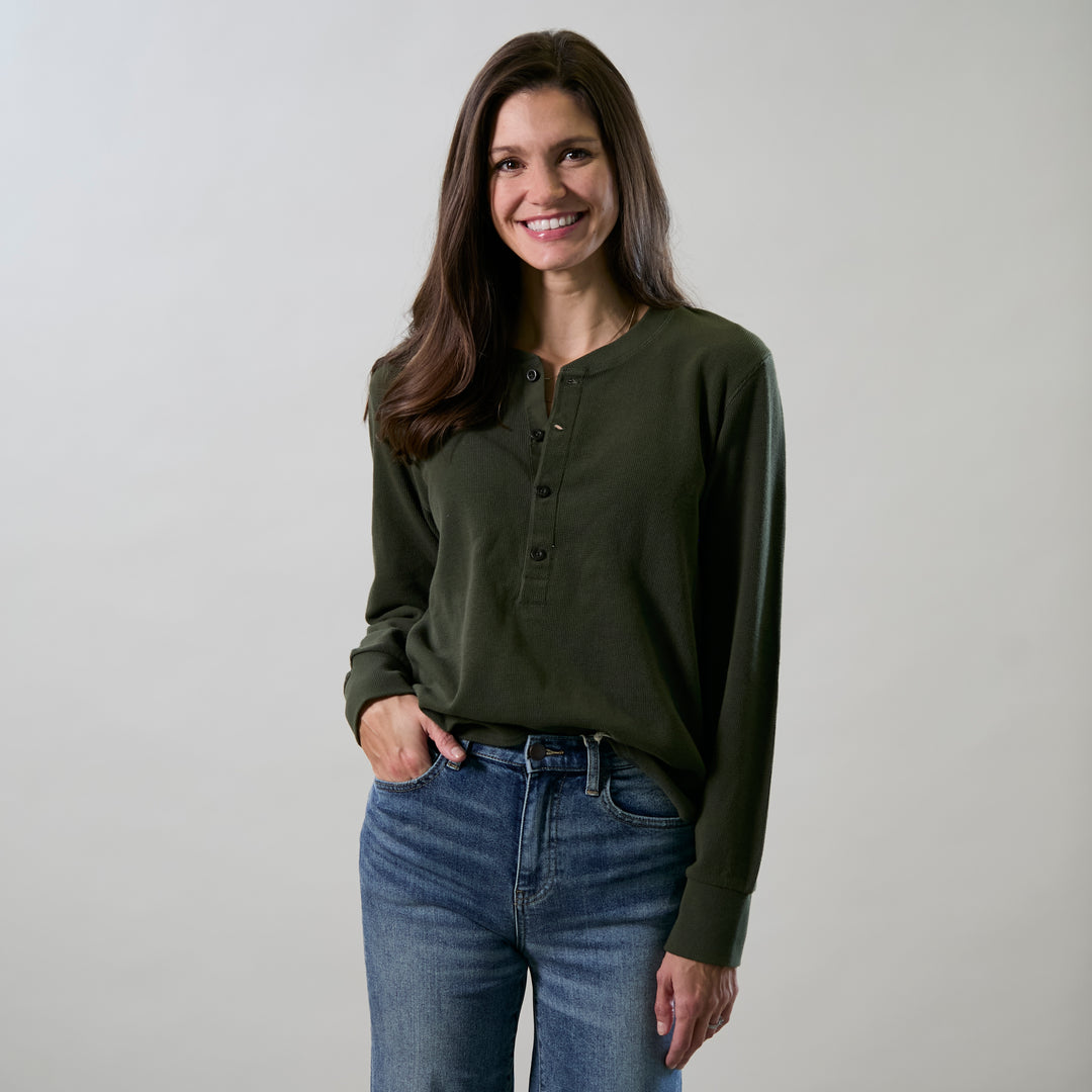A woman with long brown hair, dressed in the Women's Elko Henley- Forest Green and blue jeans, smiles with one hand in her pocket against a plain light background.