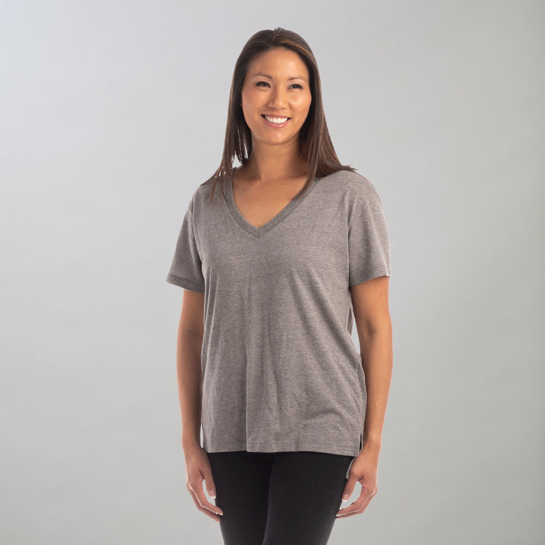 A woman with long brown hair smiles against a light gray background, wearing the Women's V-Neck Tri-Blend Tee in Charcoal Grey Heather, featuring an oversized silhouette and paired with black pants.