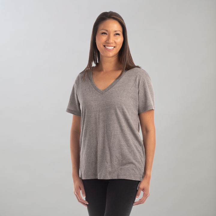 A woman with long brown hair smiles against a light gray background, wearing the Women&