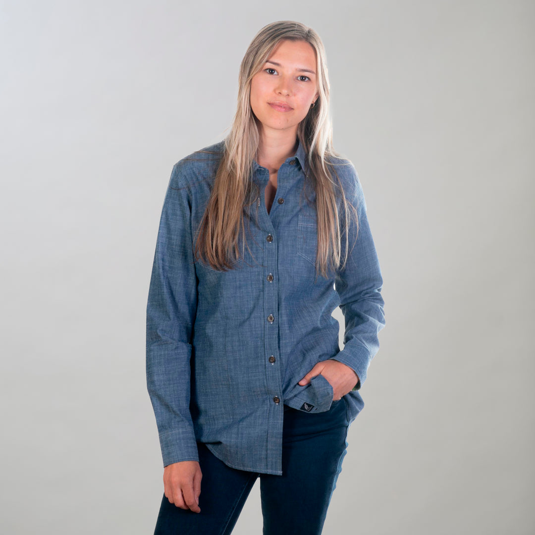 A woman with long blonde hair wears the Women's Cascade Shirt- Indigo Blue Chambray and dark pants, standing against a light gray background with one hand in her pocket and a neutral expression.
