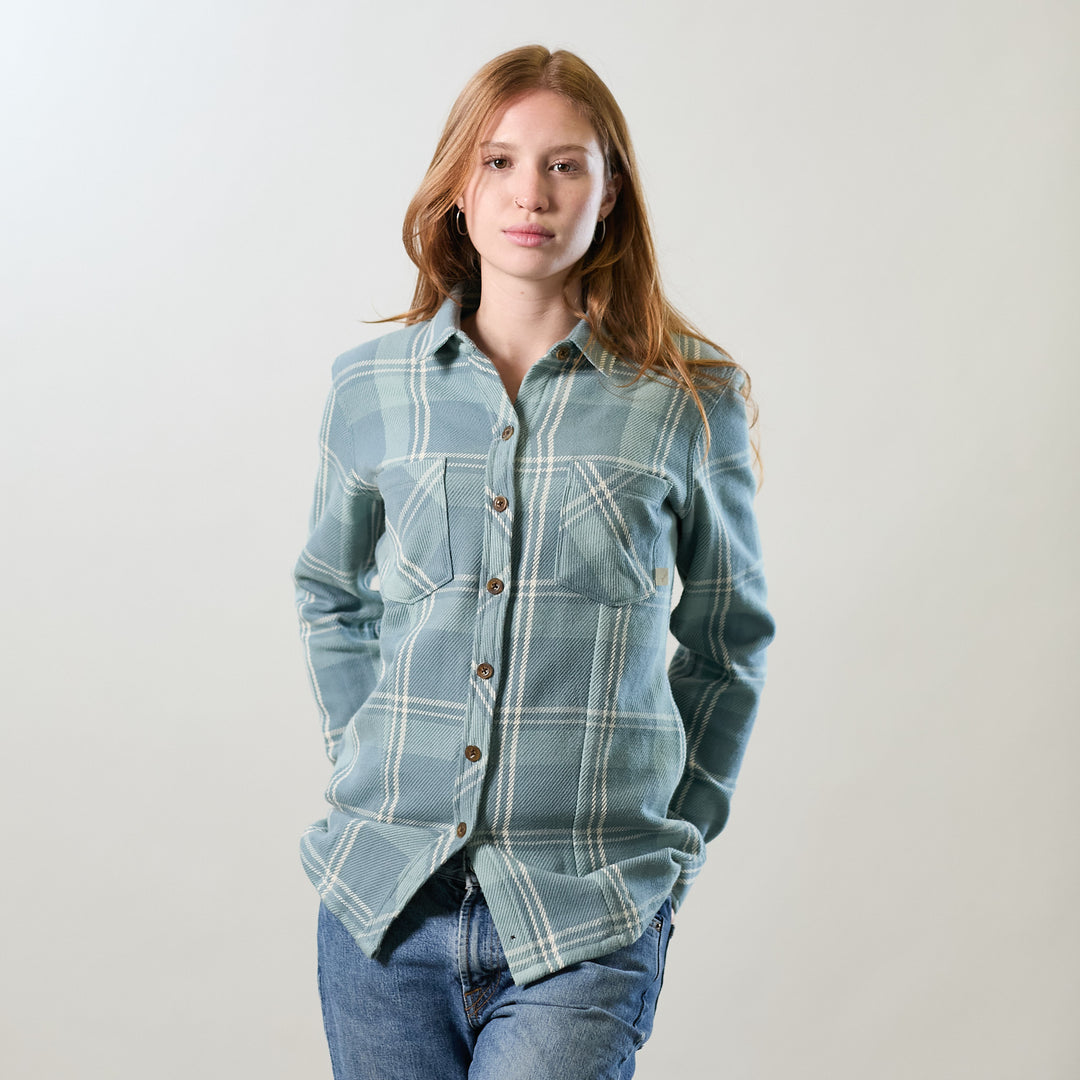 A woman with long red hair stands against a plain background, wearing the Women's Fireside Flannel in Island Blue and blue jeans, hands in her pockets with a neutral expression.