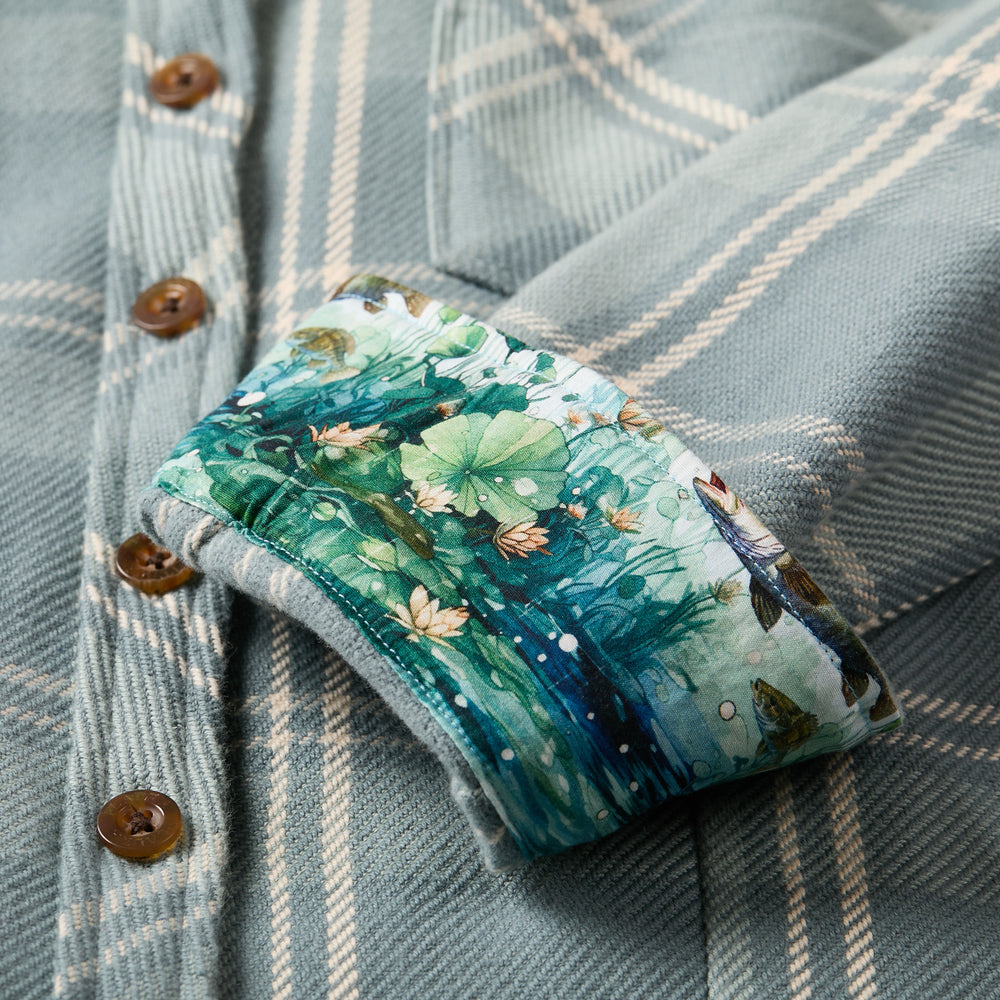 A close-up of the Women's Fireside Flannel- Island Blue shows blue and white plaid with brown buttons. The rolled cuff reveals a nature-themed lining with leaves, water lilies, and frogs—crafted from soft organic cotton flannel.