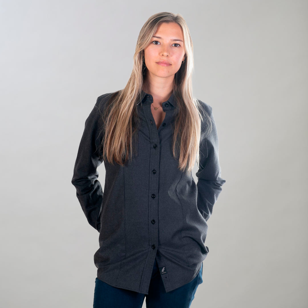 A woman with long, straight blonde hair stands facing the camera in a Women's Cascade Flannel Shirt - Jet Black Heather and dark pants, hands behind her back, against a plain light background.