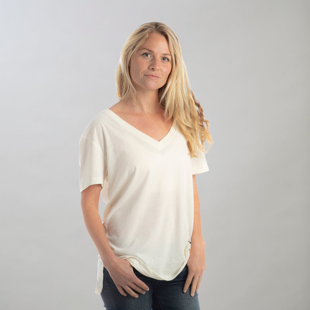 A woman with long blonde hair wears a Women's V-Neck Tri-Blend Tee in Marshmallow White Heather, styled loosely with dark jeans, standing against a light gray background with a soft expression.