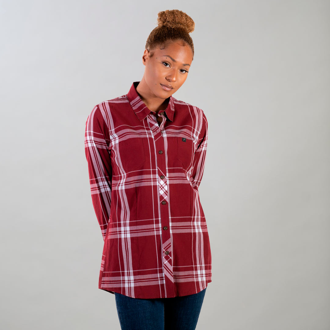A woman with curly hair in a bun wears the Women's Breeze Shirt- Merlot Red, paired with dark jeans, standing against a plain light gray background with her hands behind her back.