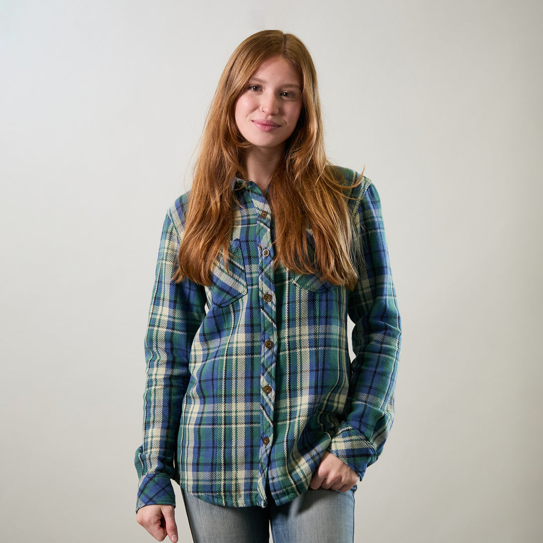 A young woman with long, reddish-brown hair smiles slightly against a plain light background, wearing the Women's Fireside Flannel in Primrose Green and light jeans, with one hand in her pocket.