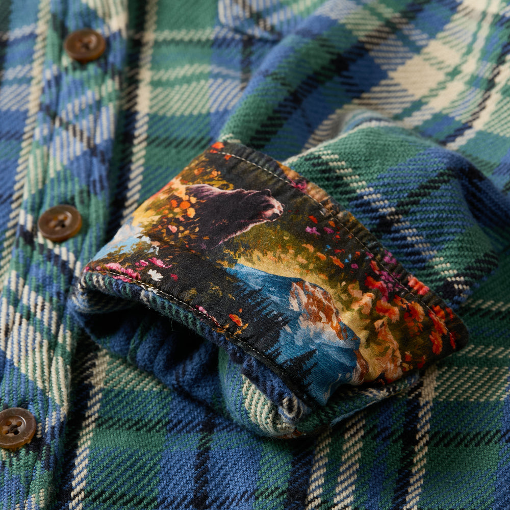A close-up of the Women's Fireside Flannel- Primrose Green shows green and blue plaid with brown buttons. The rolled-up cuff reveals a vibrant lining with a bear, trees, river, and colorful foliage nature scene.