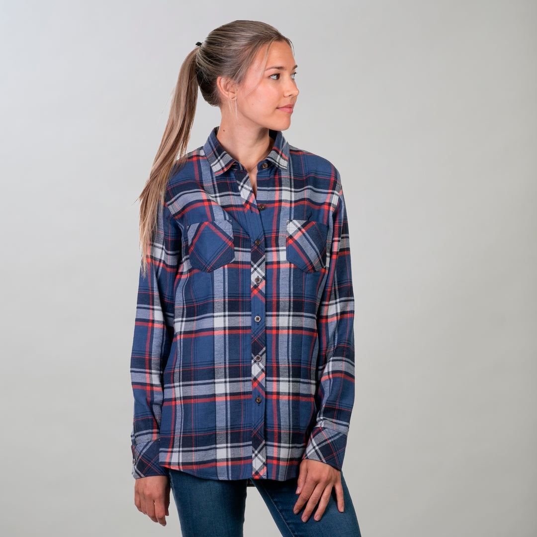 A woman with long, light brown hair in a ponytail wears the Women's Every Day Double Weave Shirt- Sunset Blue, featuring a blue, red, and white plaid pattern. She pairs it with blue jeans and stands against a plain light gray background looking left.