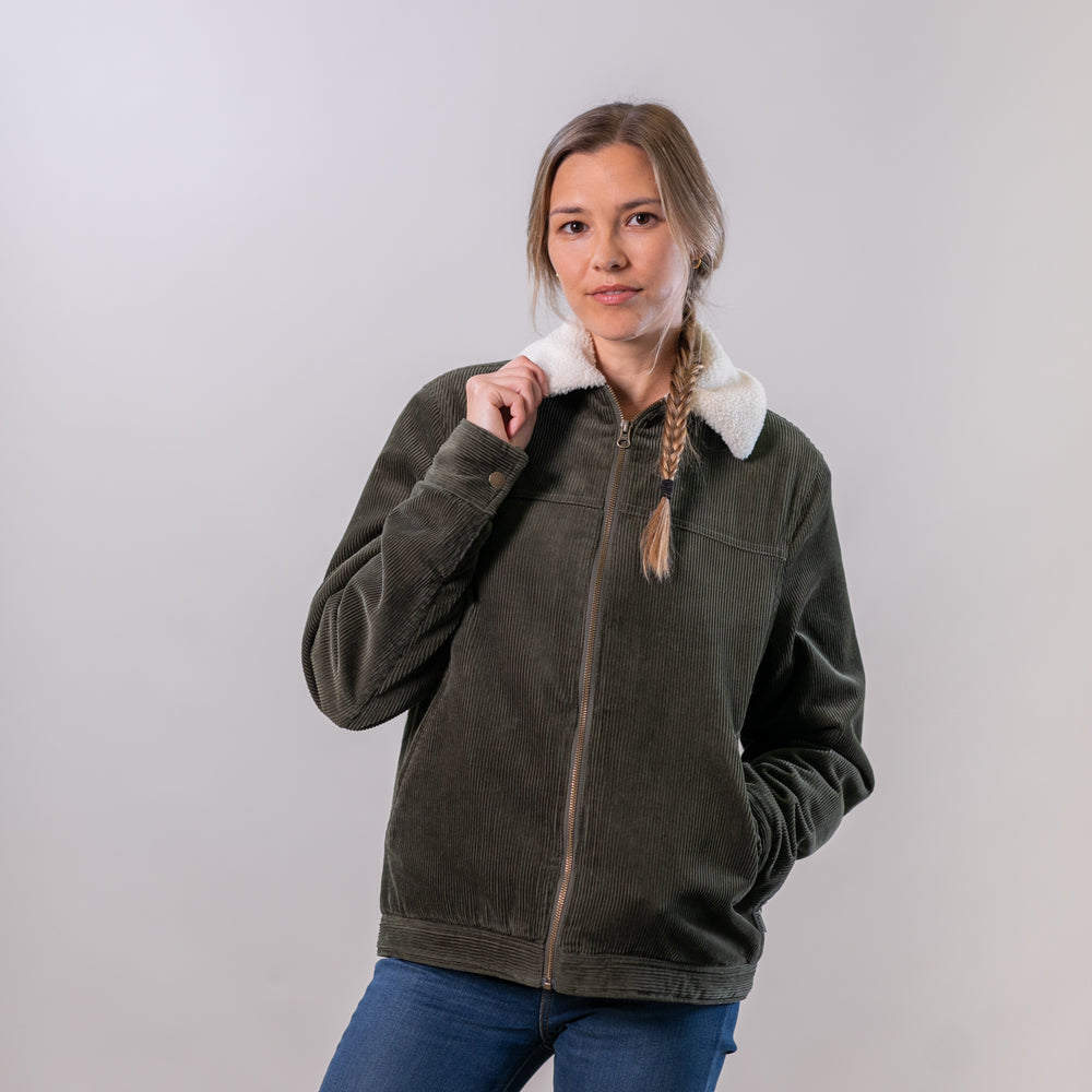 A woman with a braid poses against a light gray background, holding the collar of the Women's Stinson Jacket- Pine Green, which features a sherpa lining and white fleece collar.