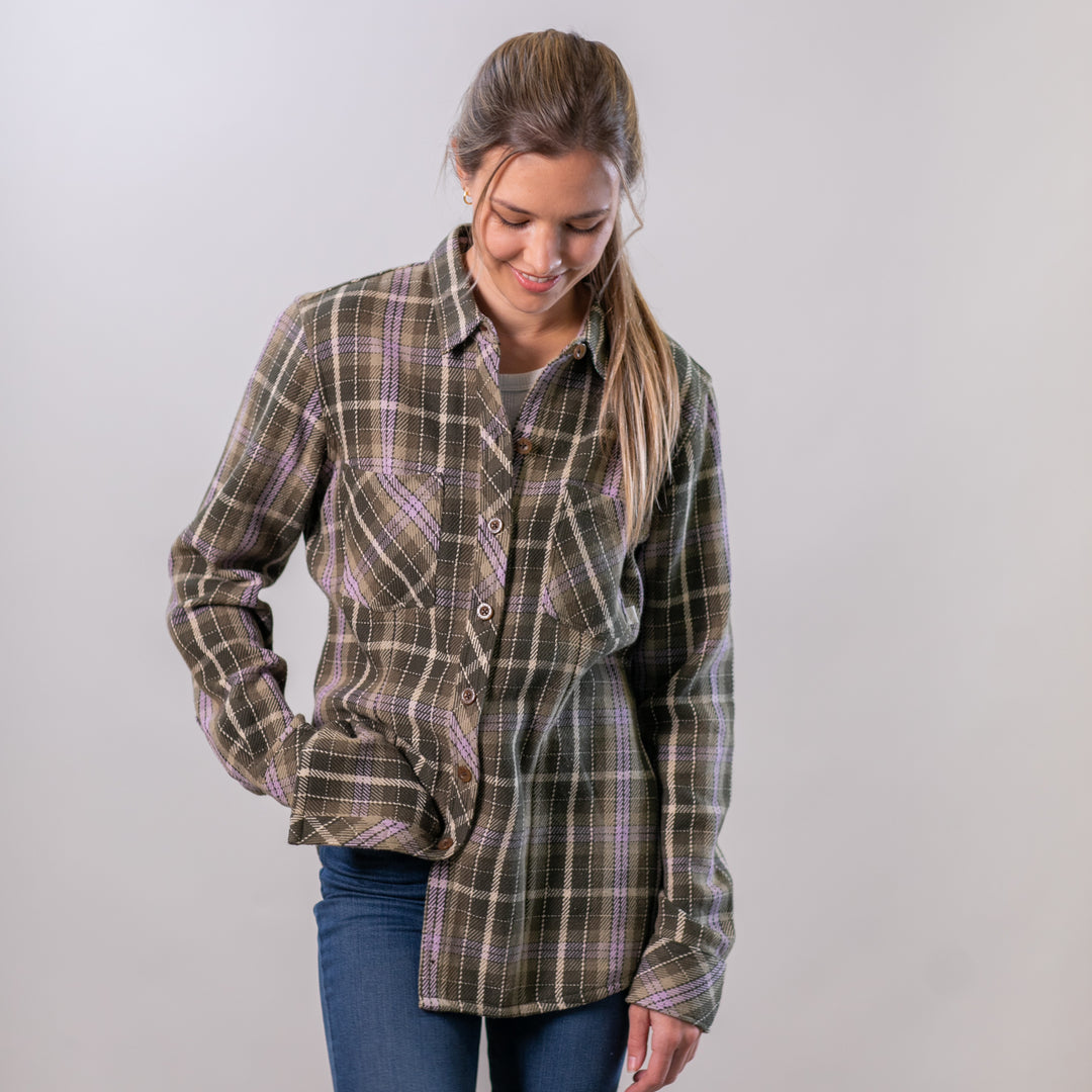 A woman with long hair in a ponytail smiles and looks down with one hand in her pocket, wearing the Women's Fireside Flannel- Grove Green and blue jeans against a plain light background.