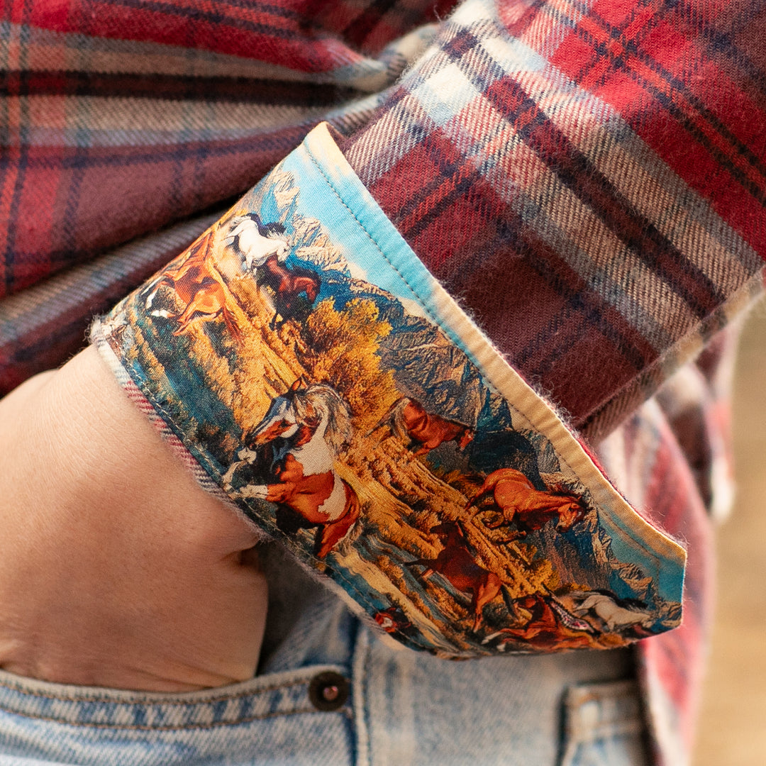A hand in a jeans pocket wears the Pladra Women's Every Day Flannel Shirt–Harvest Red with wild horses on the cuff.
