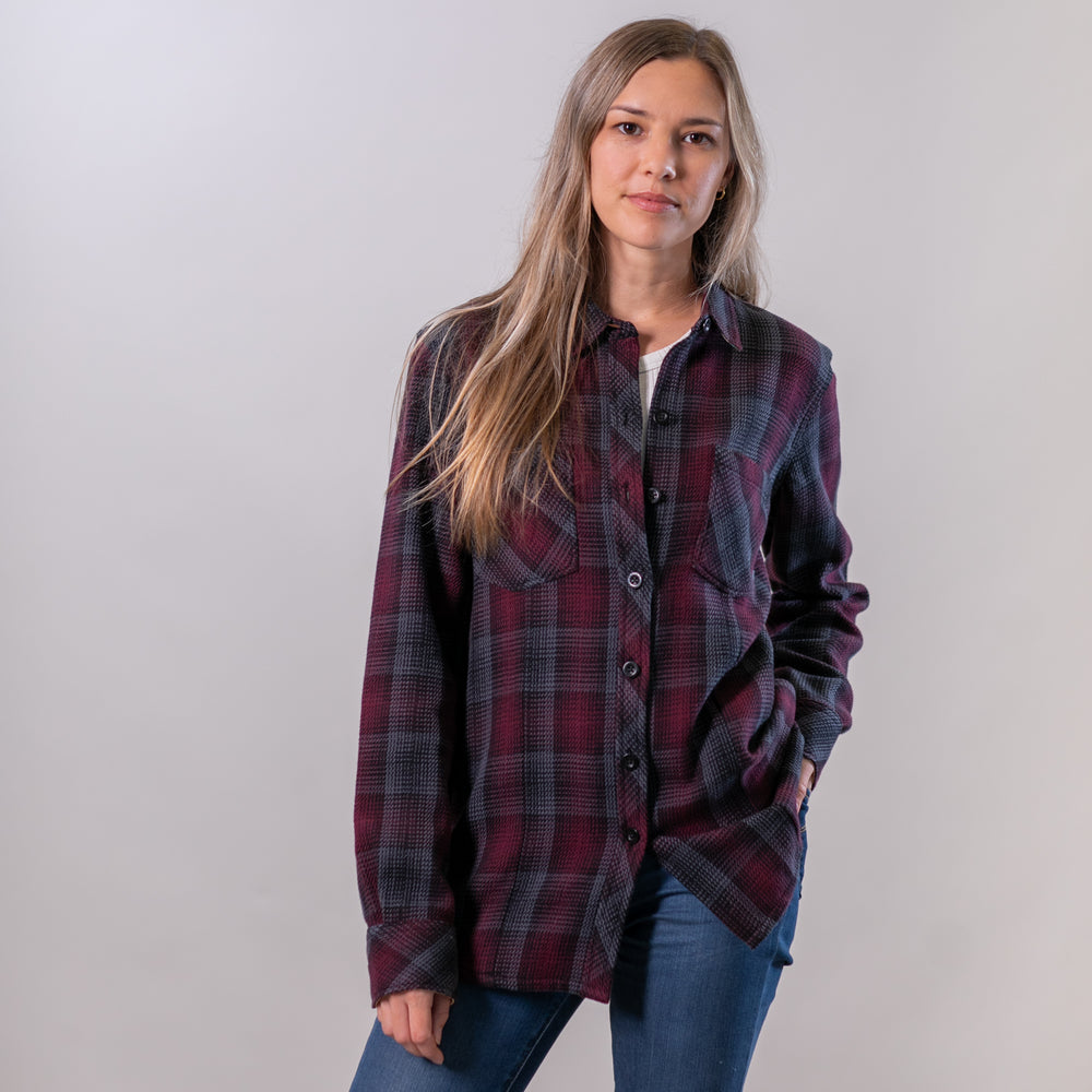 A woman with long brown hair wears the Women's Fireside Flannel - Hickory Red and blue jeans, standing against a plain light background with one hand in her pocket and a neutral expression.