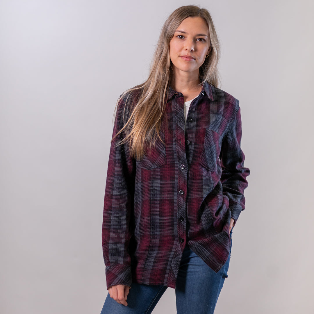 A woman with long brown hair wears the Women's Fireside Flannel - Hickory Red and blue jeans, standing against a plain light background with one hand in her pocket and a neutral expression.