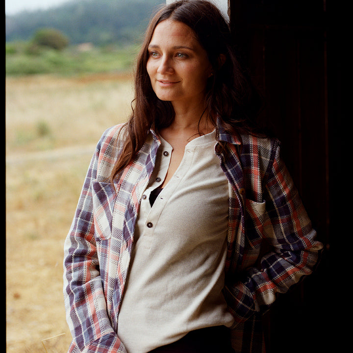 Woman wearing a plaid shirt over a white top, standing outdoors with a field and trees in the background.