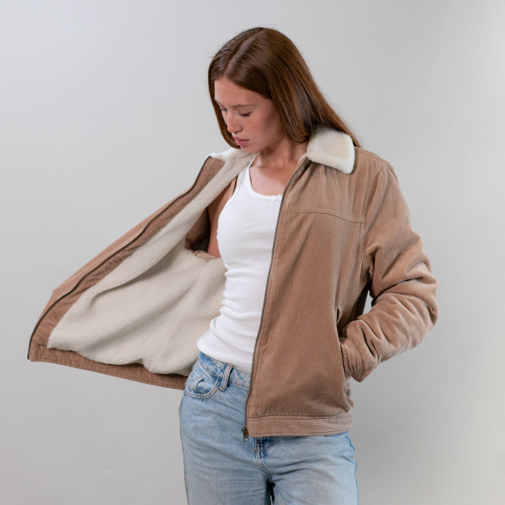 A woman in jeans and a white top opens a Pladra Women's Stinson Jacket in Oak Tan, admiring the cozy lining.