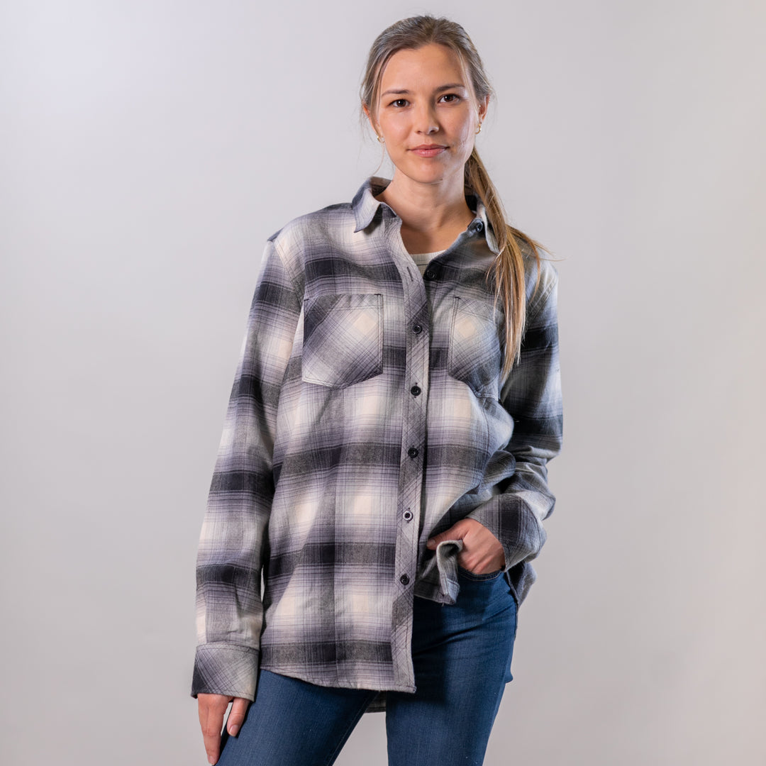A woman with light brown hair in a ponytail wears the Women's Every Day Flannel Shirt in Sterling Grey Heather and blue jeans, standing against a plain light background with her hand in her pocket and a slight smile.