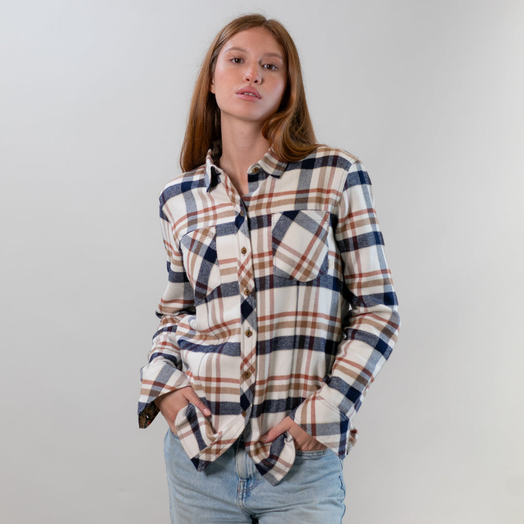 A woman with long brown hair models the Women's Every Day Elite Flannel Shirt - Whitefish Tan, featuring white, brown, and blue hues in premium cotton. She pairs it with light blue jeans and stands confidently with hands in her pockets against a plain background.