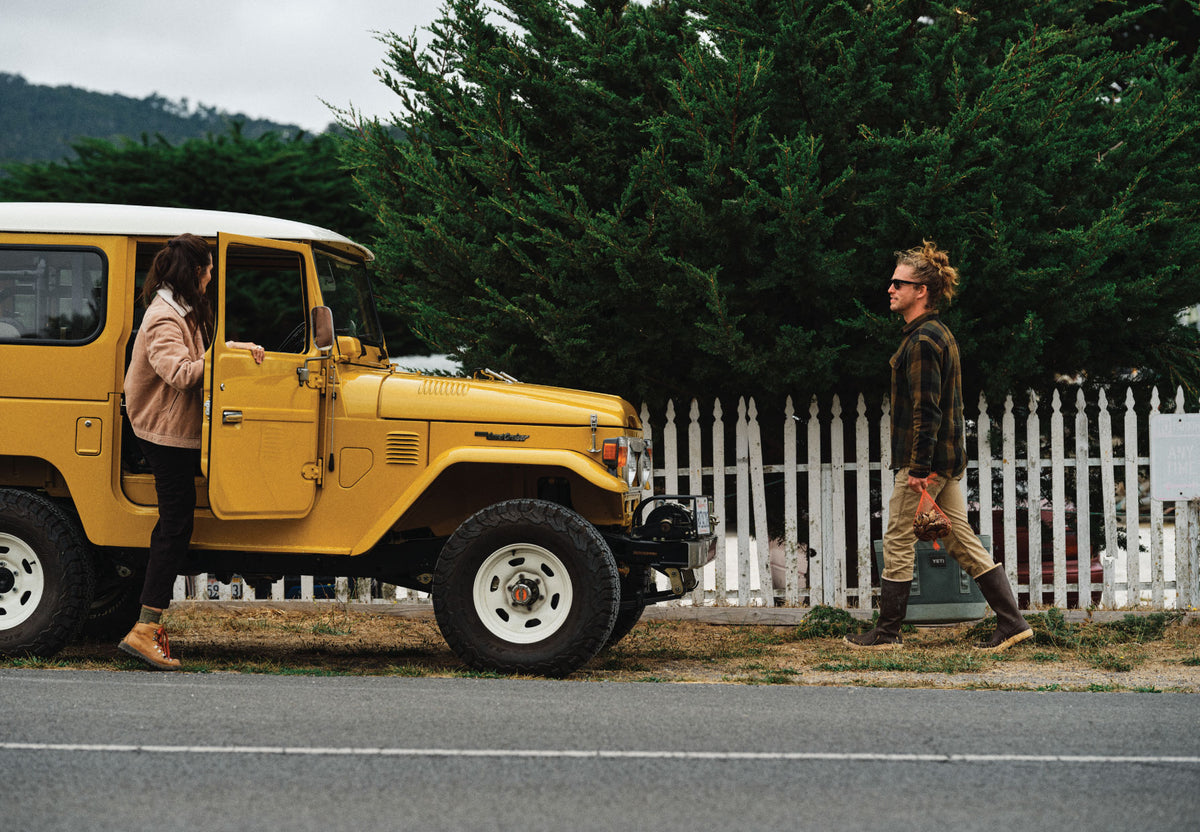 A person steps out of a yellow SUV while another person walks a dog by a white picket fence and trees.