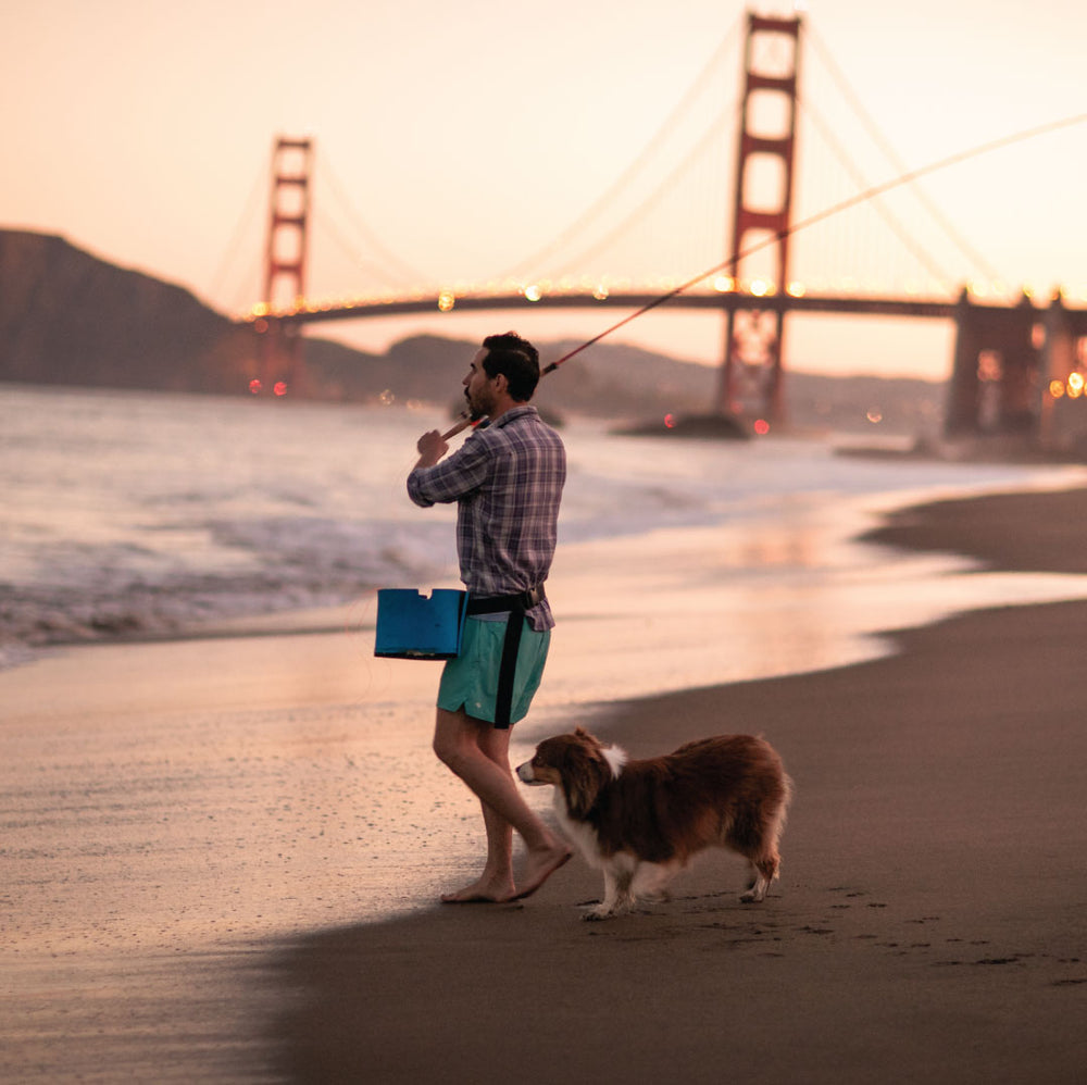 A man fishing barefoot on the beach at sunset with his dog, holding a blue tackle box and wearing the Men's Every Day Elite Flannel Shirt- Geary Grey. The Golden Gate Bridge appears in the background.