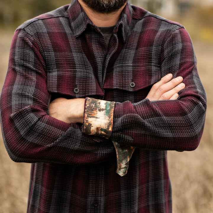 A bearded man stands outdoors with his arms crossed, wearing the Men&