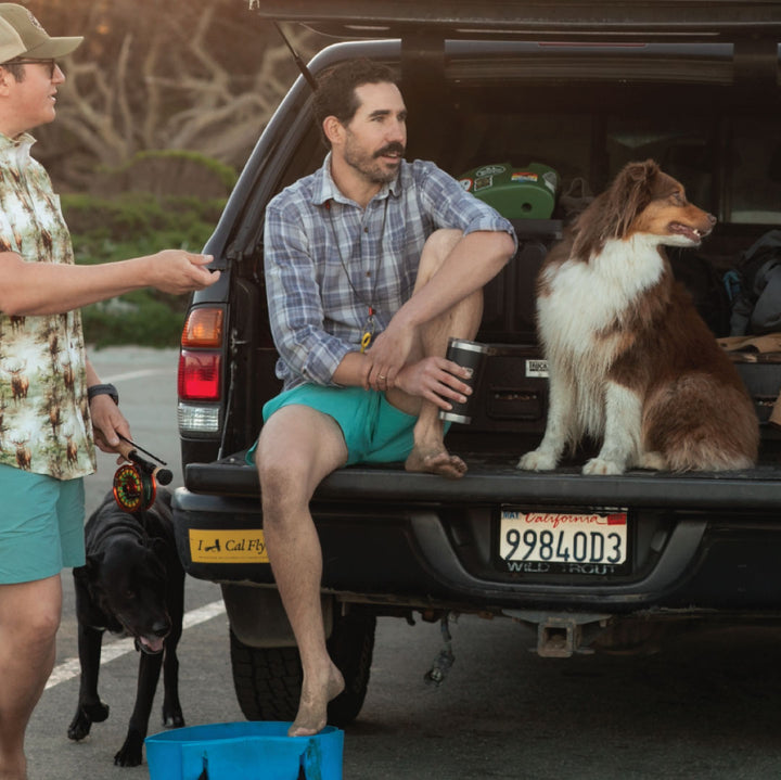 Two men and two dogs stand by an open SUV trunk. One man, barefoot and holding a cup, sits with a brown and white dog. The other, in the Men&