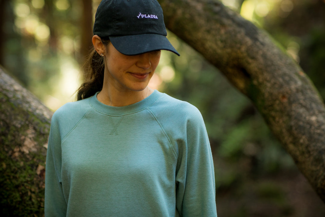 A woman stands outdoors in a forest, sunlight filtering through the trees, wearing a black cap and the Women&