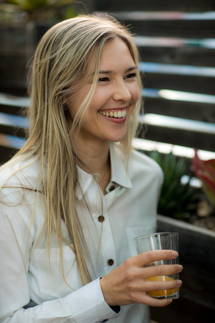 Woman in Pladra Women’s Breeze Shirt–Whisper Grey enjoys a drink outdoors, surrounded by plants.