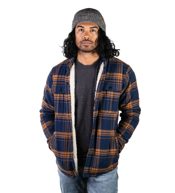 A man with long curly hair in a gray beanie, dark t-shirt, and jeans stands against a white background wearing the Men&
