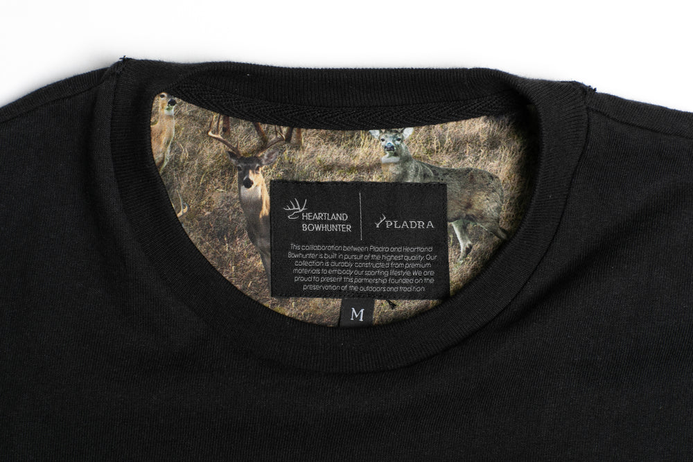 Close-up of the Men's Heavyweight Pocket Tee- HB Onyx Black collar, featuring a printed deer scene on the inner lining and a label reading Heartland Bowhunter x Pladra with additional text below.