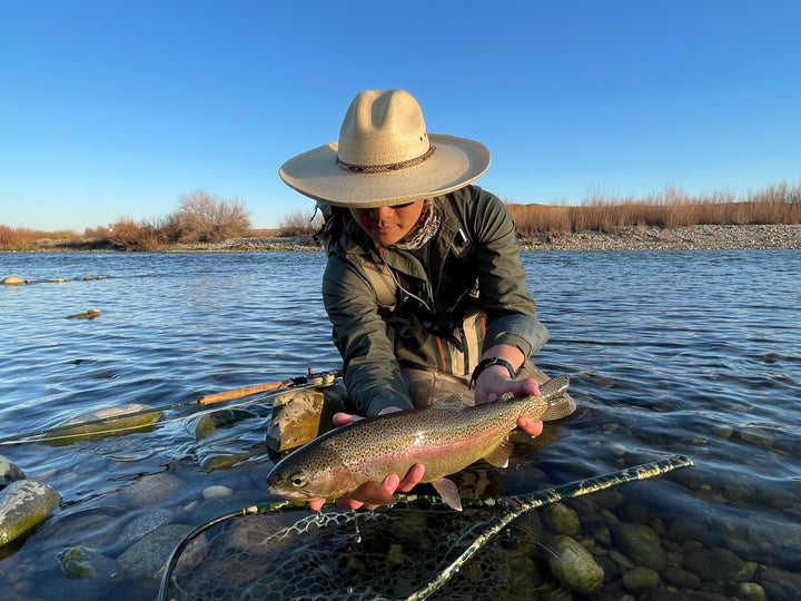 Man in hat holds a trout over shallow river, wearing Pladra Men&