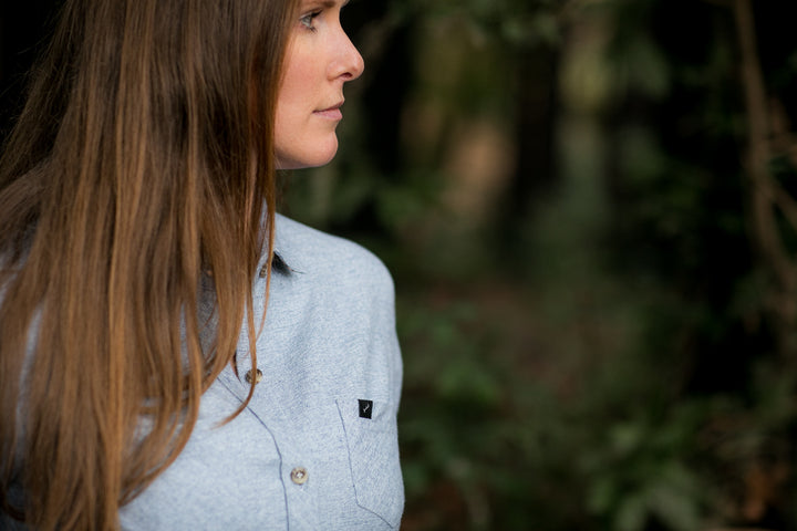 A woman with long brown hair stands outdoors amid trees, wearing the Women&
