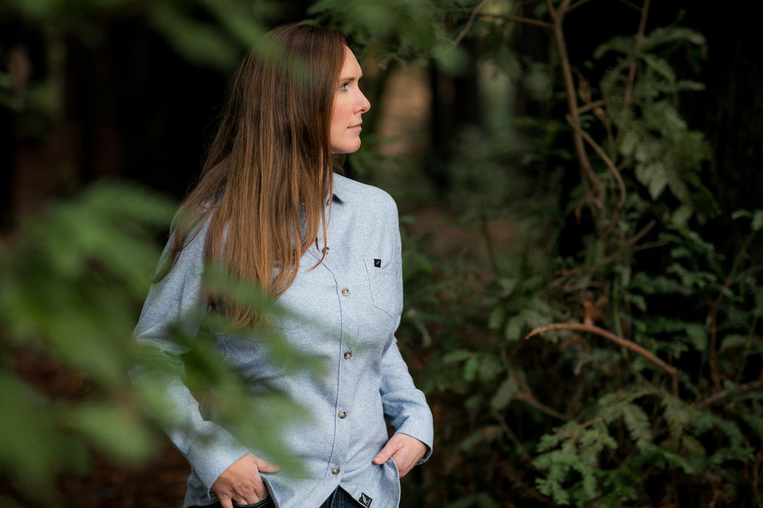 A woman with long brown hair stands outdoors among green trees, wearing the Women&