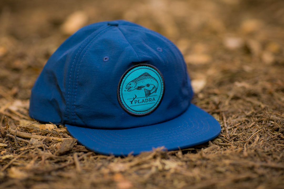 The Nylon Trout Hat- Navy Blue features a circular patch with an illustrated fish and "PLADRA" text. Its quick-drying design makes it an ideal addition to any fishing hat collection. Shown resting on brown mulch.