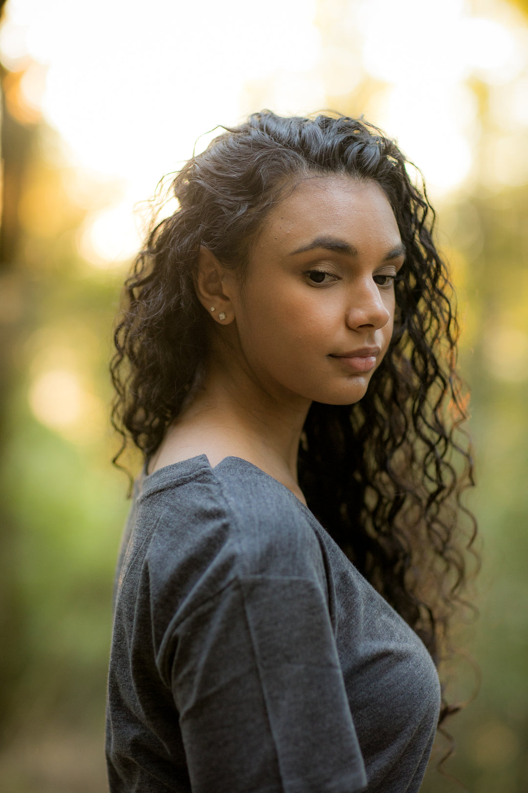 A young woman with long curly hair stands outdoors in soft sunlight, wearing a Women&