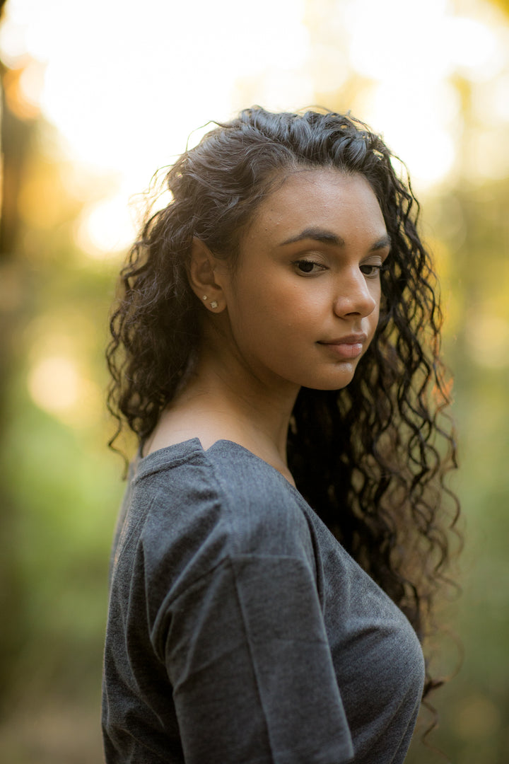 A young woman with long curly hair stands outdoors in soft sunlight, wearing a Women&