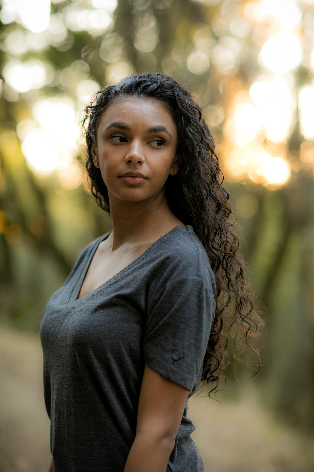 A woman with long curly hair wears a Women’s V-Neck Tri-Blend Tee in Charcoal Grey Heather, standing outdoors and looking to the side, with trees and warm sunlight blurred in the background.