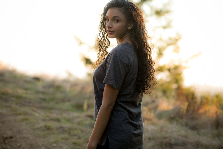 A young woman with long curly hair stands on a grassy hill at sunset, wearing the Women’s V-Neck Tri-Blend Tee in Charcoal Grey Heather and looking over her shoulder toward the camera.