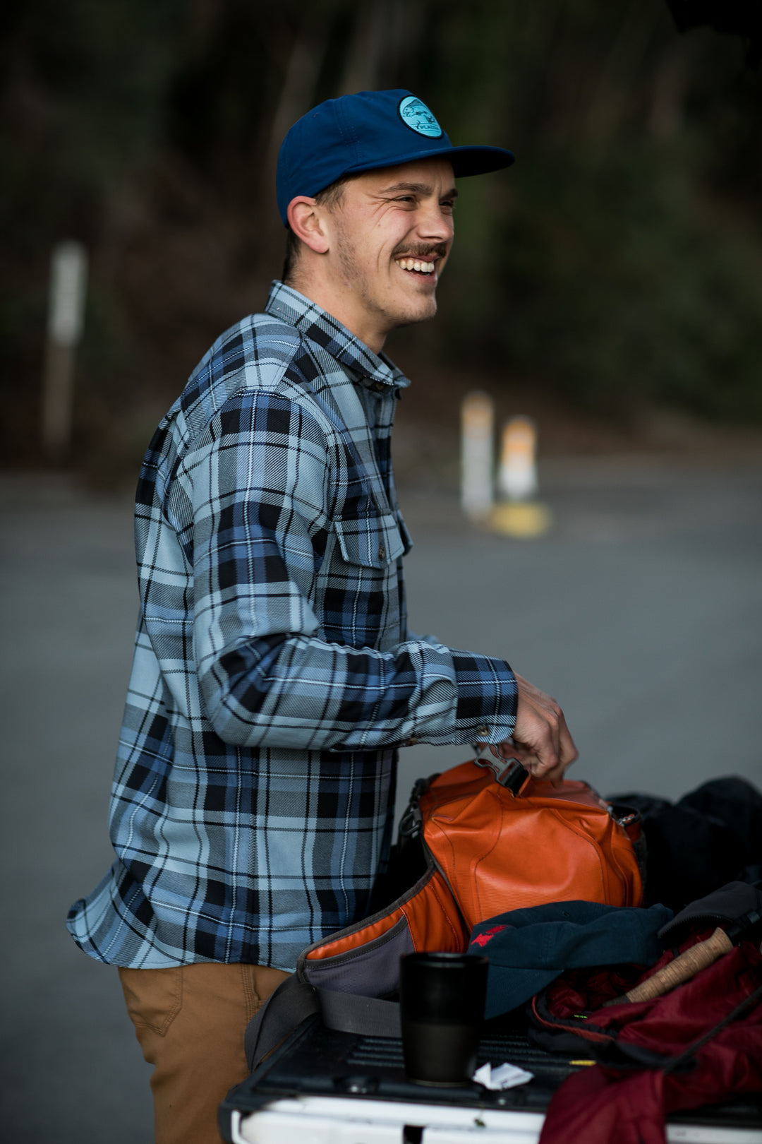 A man in a blue plaid shirt and the Nylon Trout Hat- Navy Blue smiles outdoors by a car trunk, holding an orange bag with various items. Trees and blurred road signs appear in the background.