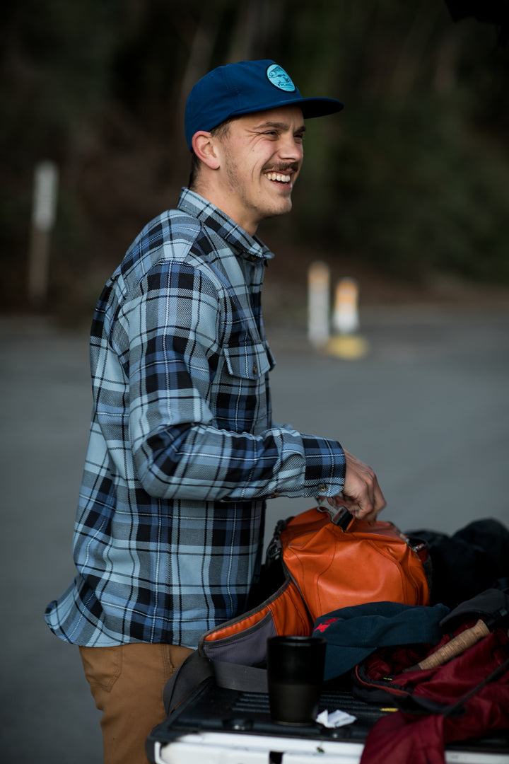 A man in a blue plaid shirt and the Nylon Trout Hat- Navy Blue smiles outdoors by a car trunk, holding an orange bag with various items. Trees and blurred road signs appear in the background.