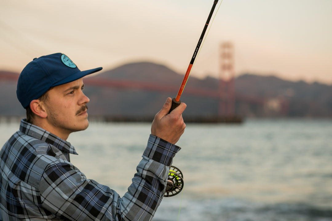 A man wearing the Nylon Trout Hat- Navy Blue is fly fishing by the water, with the Golden Gate Bridge and hills softly blurred in the background.