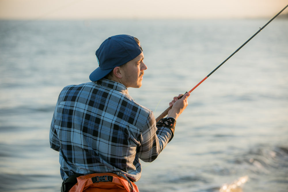 Wearing a Nylon Trout Hat in navy blue and a blue plaid shirt, a person fishes by the water, holding a rod and facing the softly sunlit scene.