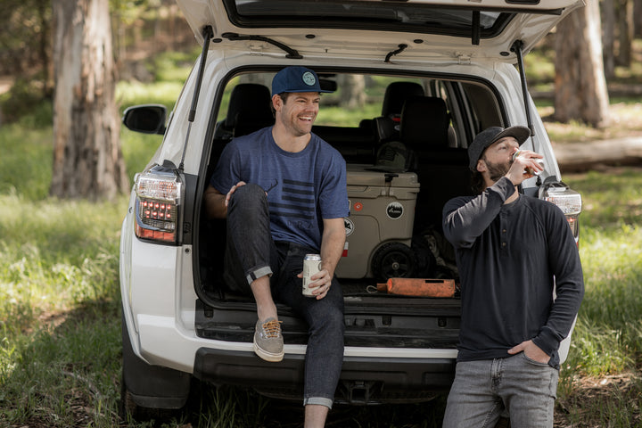 Two men enjoy the outdoors by an open SUV in the woods; one sits, holding a drink and smiling, while the other stands nearby, sipping from a can and wearing the Men&