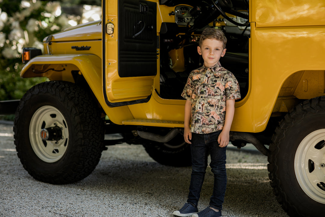A young boy in jeans and blue shoes wears a Pladra Toddler Cubs Outdoor Aloha Shirt- Mountain Lions by a yellow vintage SUV.